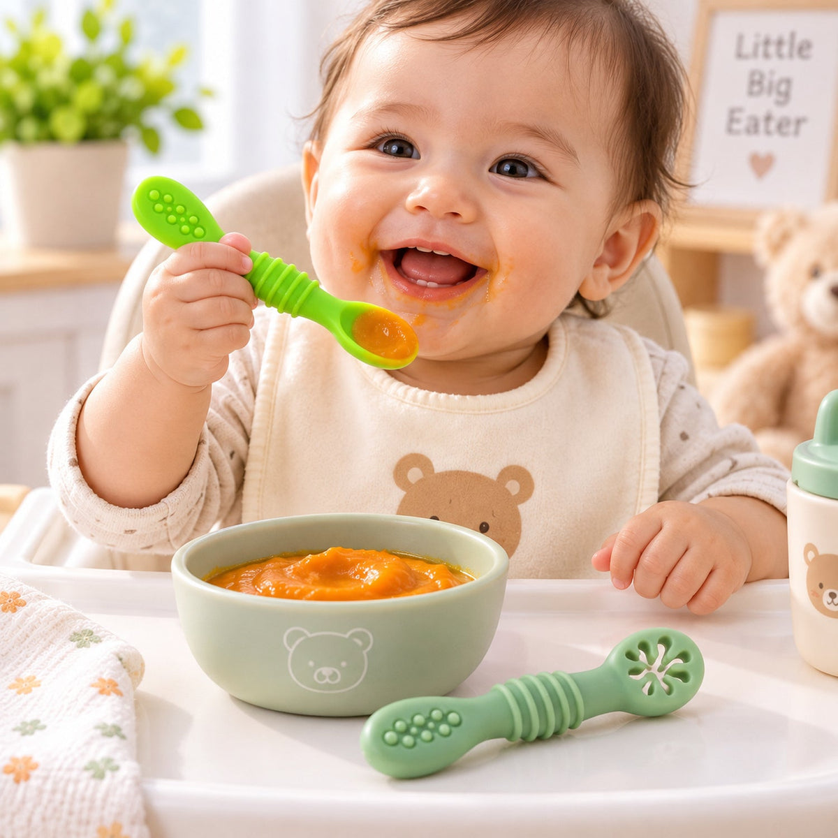 A smiling baby in a high chair holding a green silicone spoon, with a bowl of pureed food and another spoon on the tray.