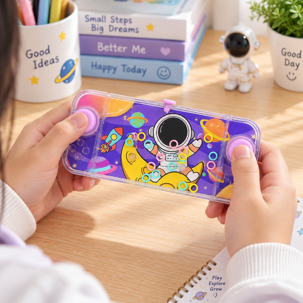 A child's hands hold a clear purple water game with an astronaut, moon, and planets design, featuring pink buttons.
