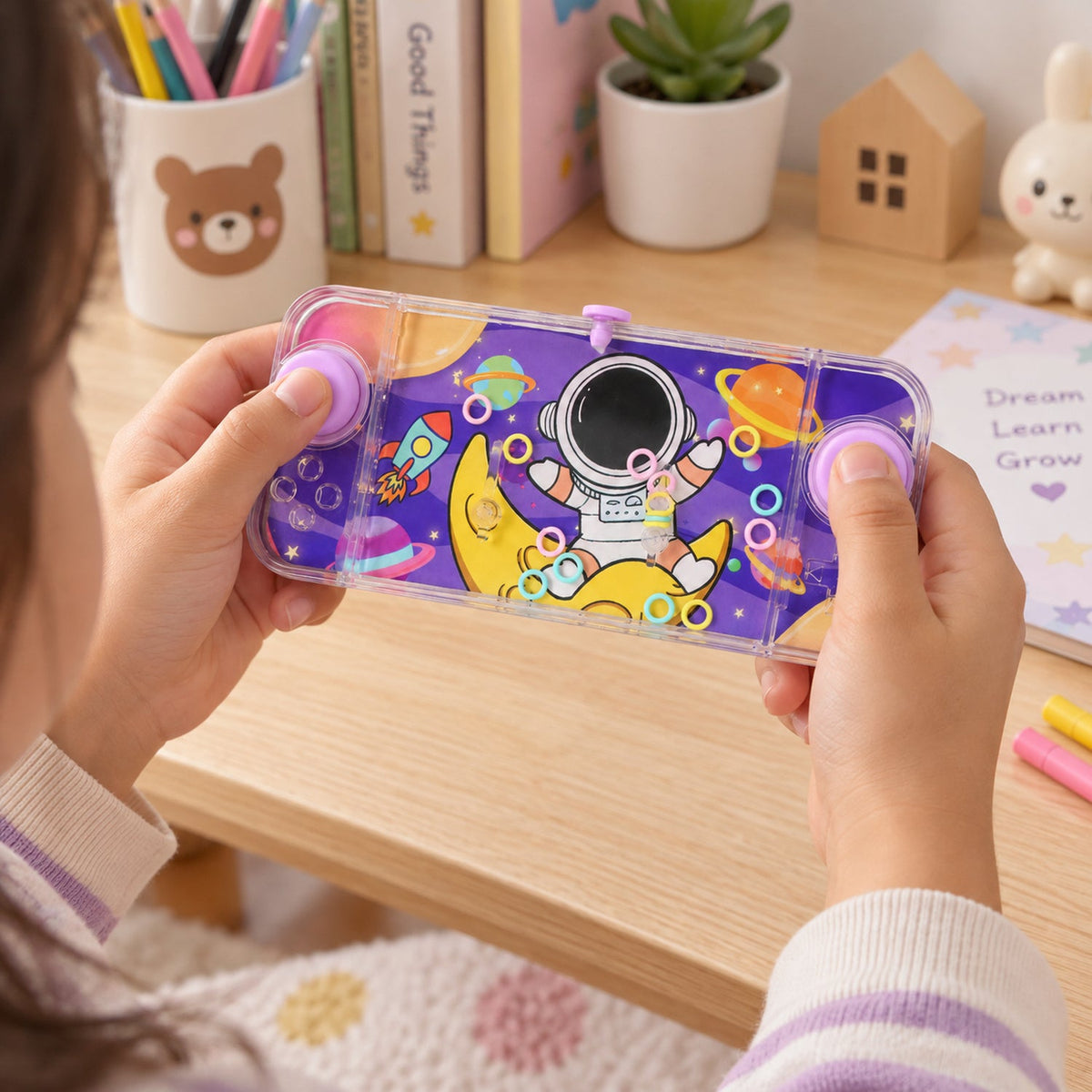 Overhead view of a child's hands holding the space-themed water game at a wooden desk with school supplies.