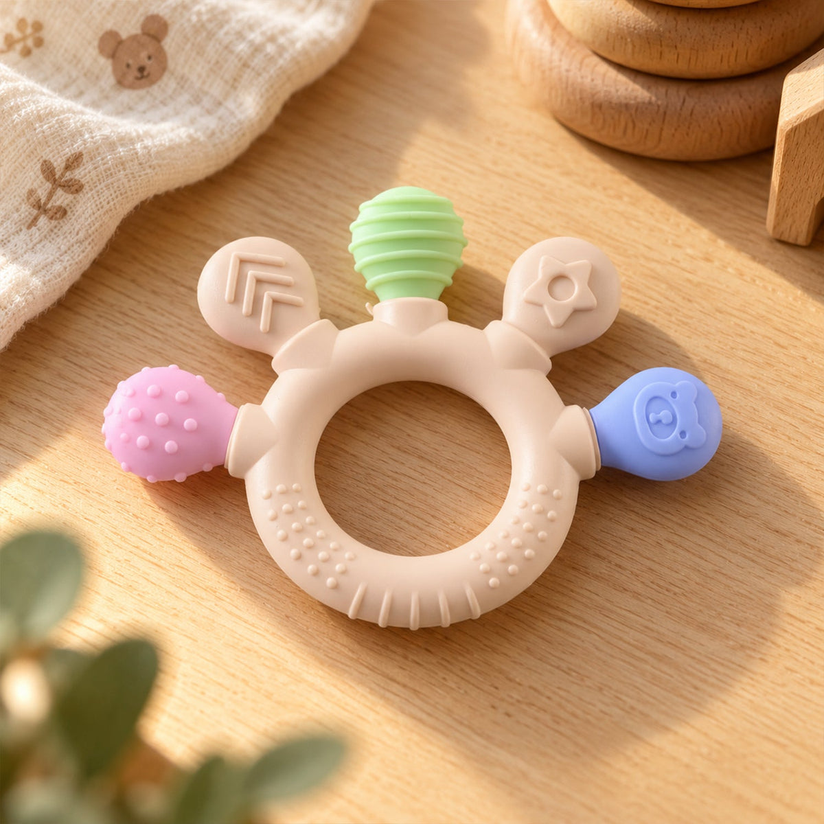 A beige teether with green, pink, and blue textured ends rests on a wooden table beside a white cloth and wooden toys.