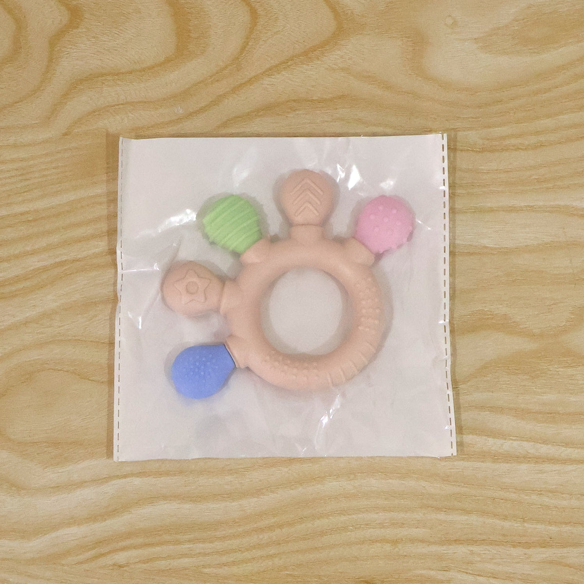 A beige teether with green, pink, and blue textured ends is sealed in a clear plastic bag on a wooden surface.
