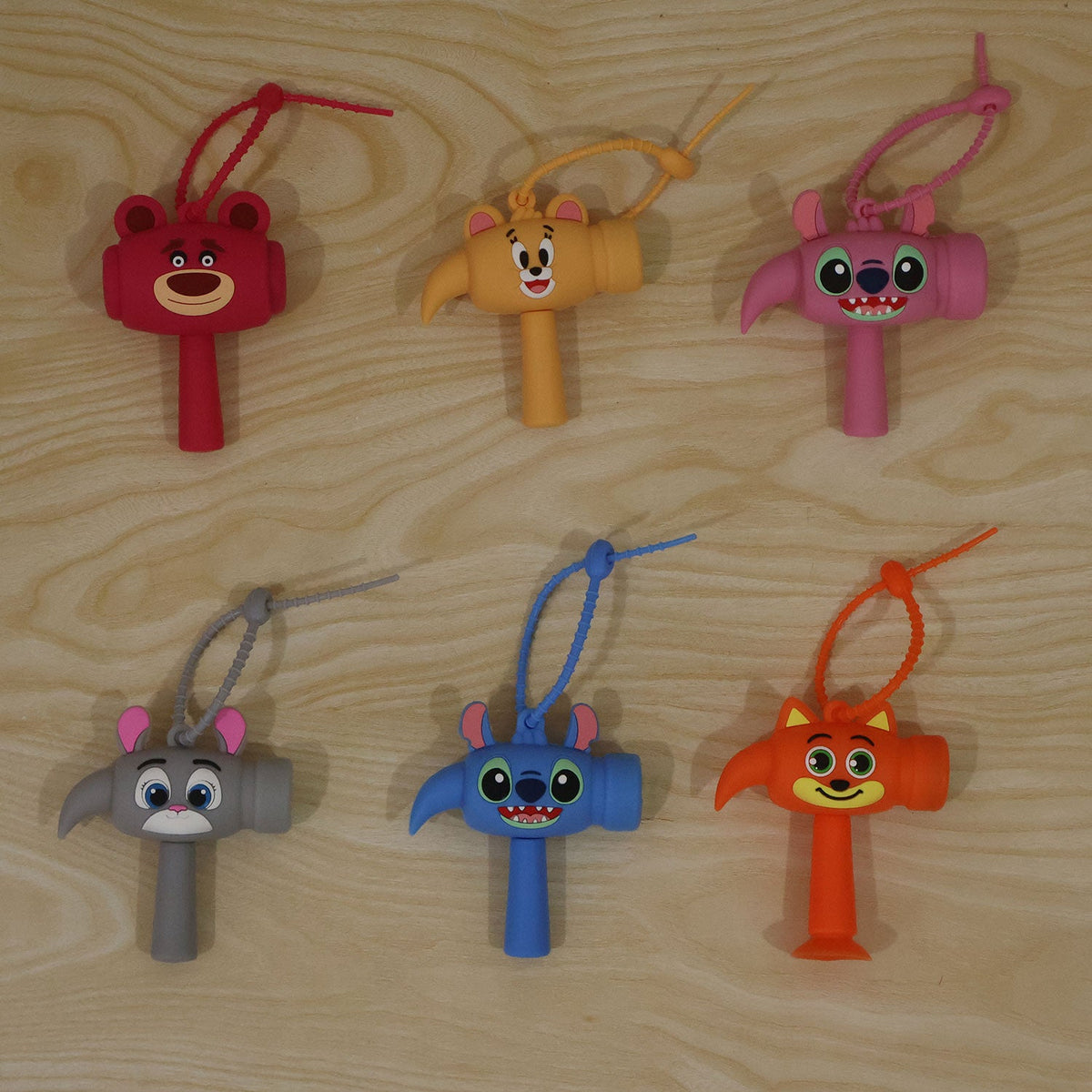 Six colorful cartoon character hammer keychains, each with a hanging loop, arranged on a light wooden surface.