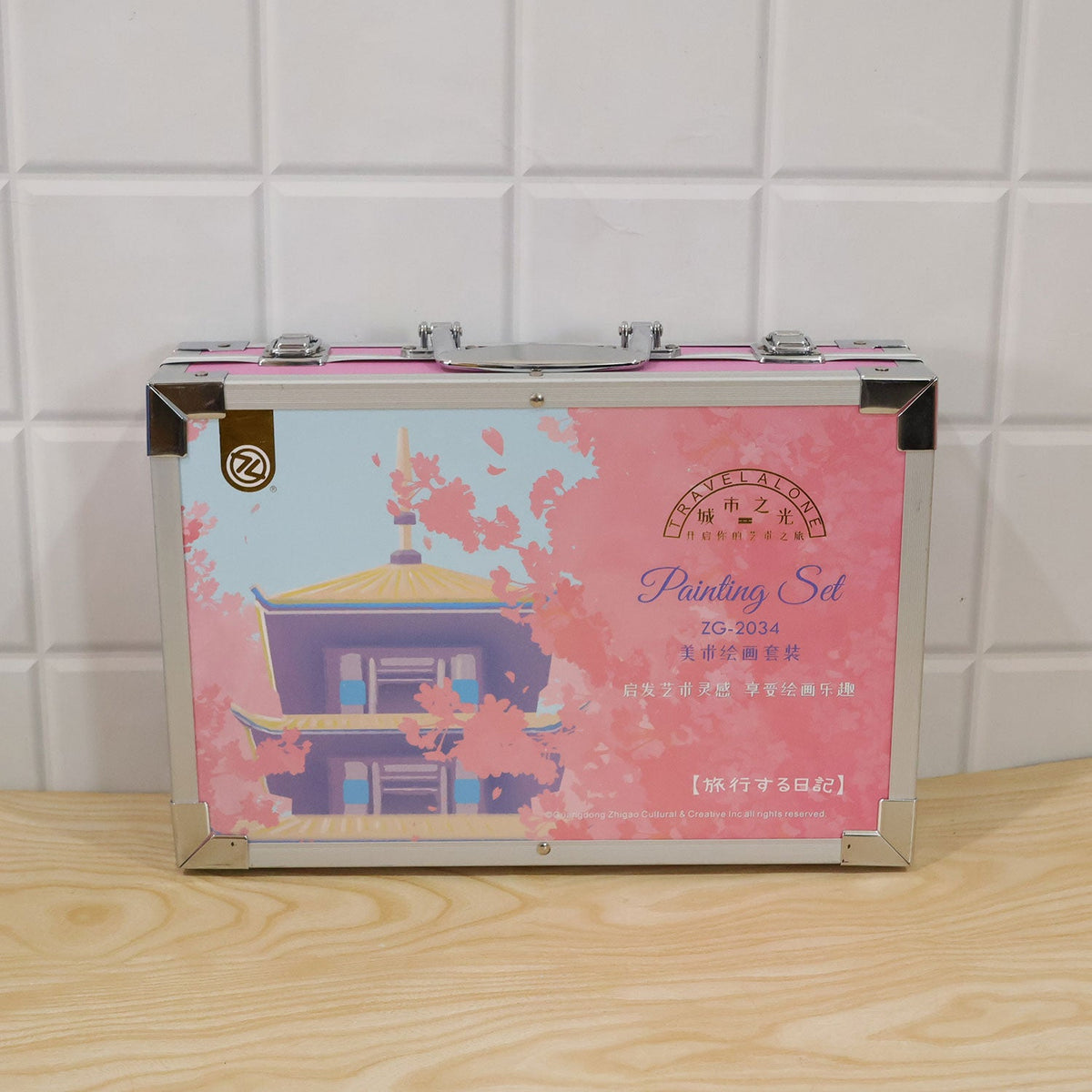 A closed pink metal art case with a pagoda and cherry blossom design rests on a light wooden surface.
