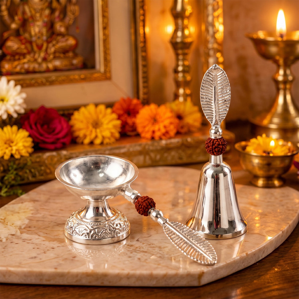 A silver finish pooja bell and diya set with leaf handles and rudraksha beads, placed on a marble slab with religious items.