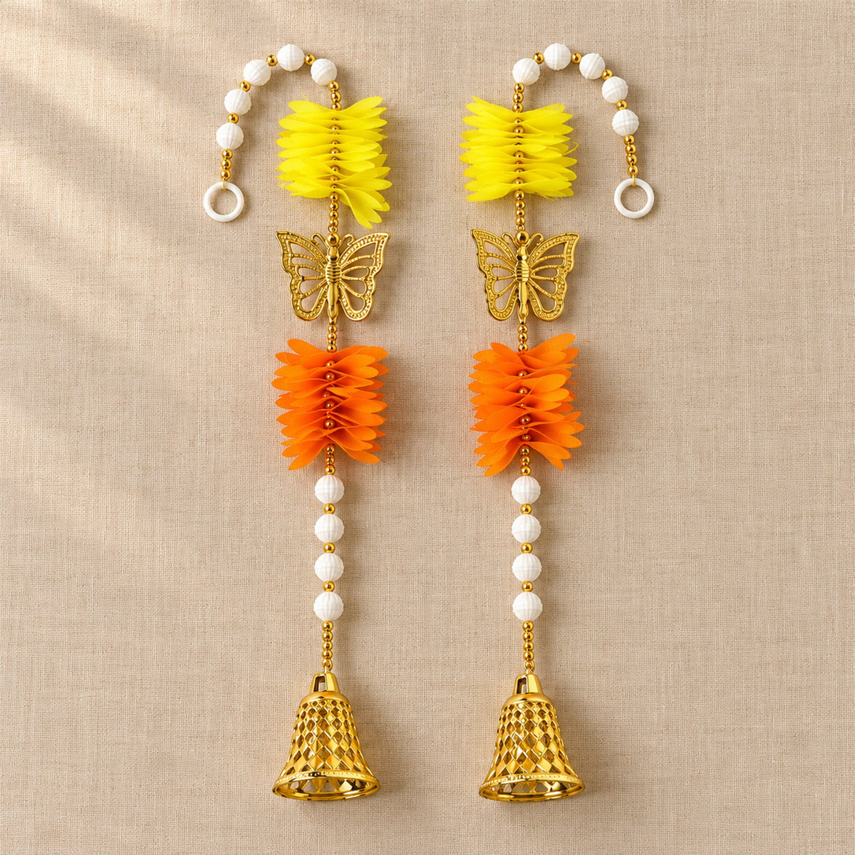 Two ornate door hangings, each with yellow and orange paper flowers, a golden butterfly, and a bell, laid flat on a textured