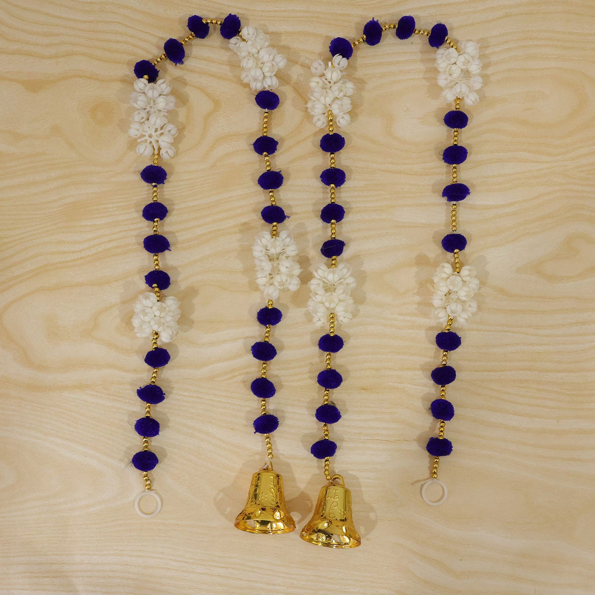 Four decorative strands, each with blue pom-poms, white floral clusters, and a golden bell, are arranged on a light wooden