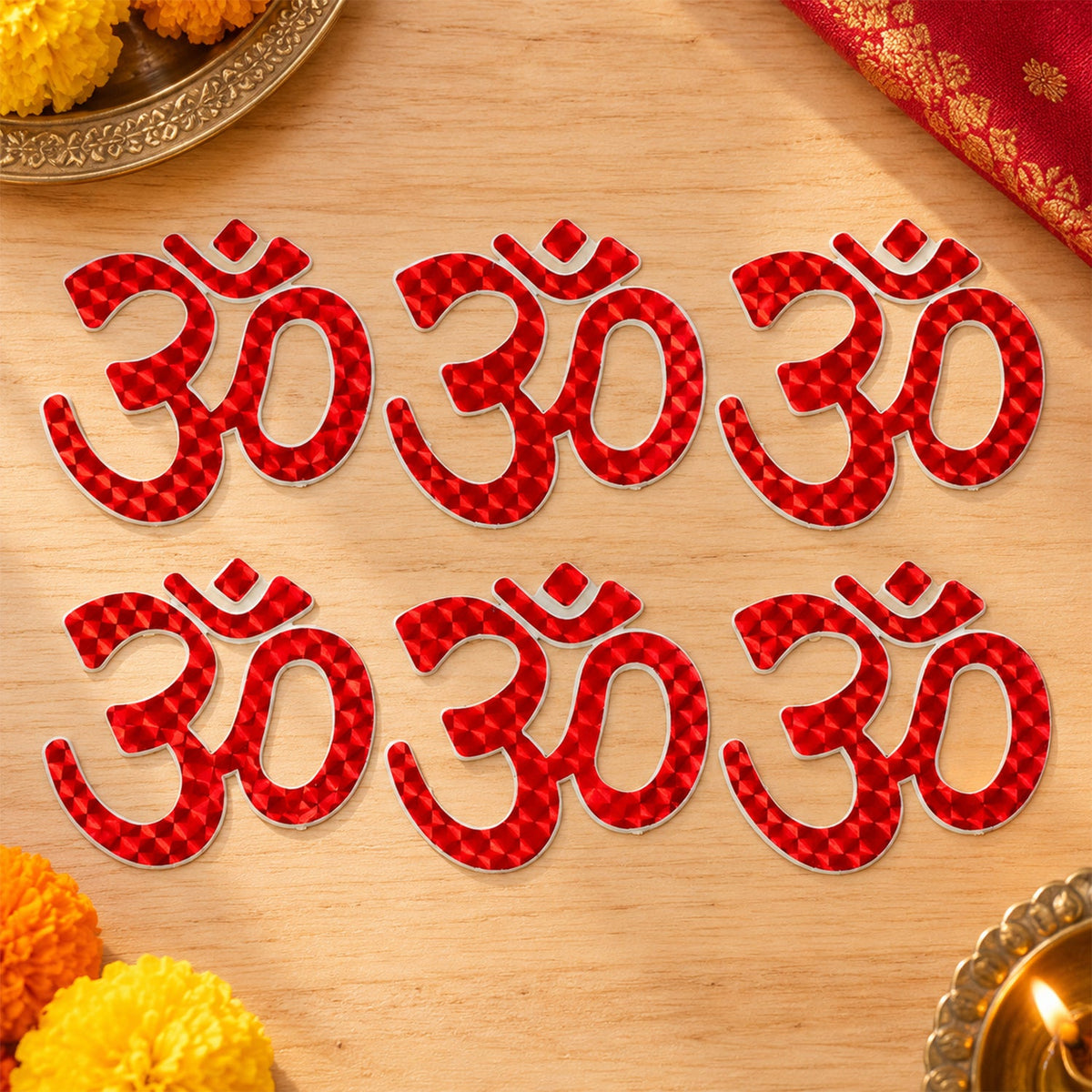 Six red Om symbol stickers are arranged in two rows on a light wooden surface, surrounded by marigold petals.
