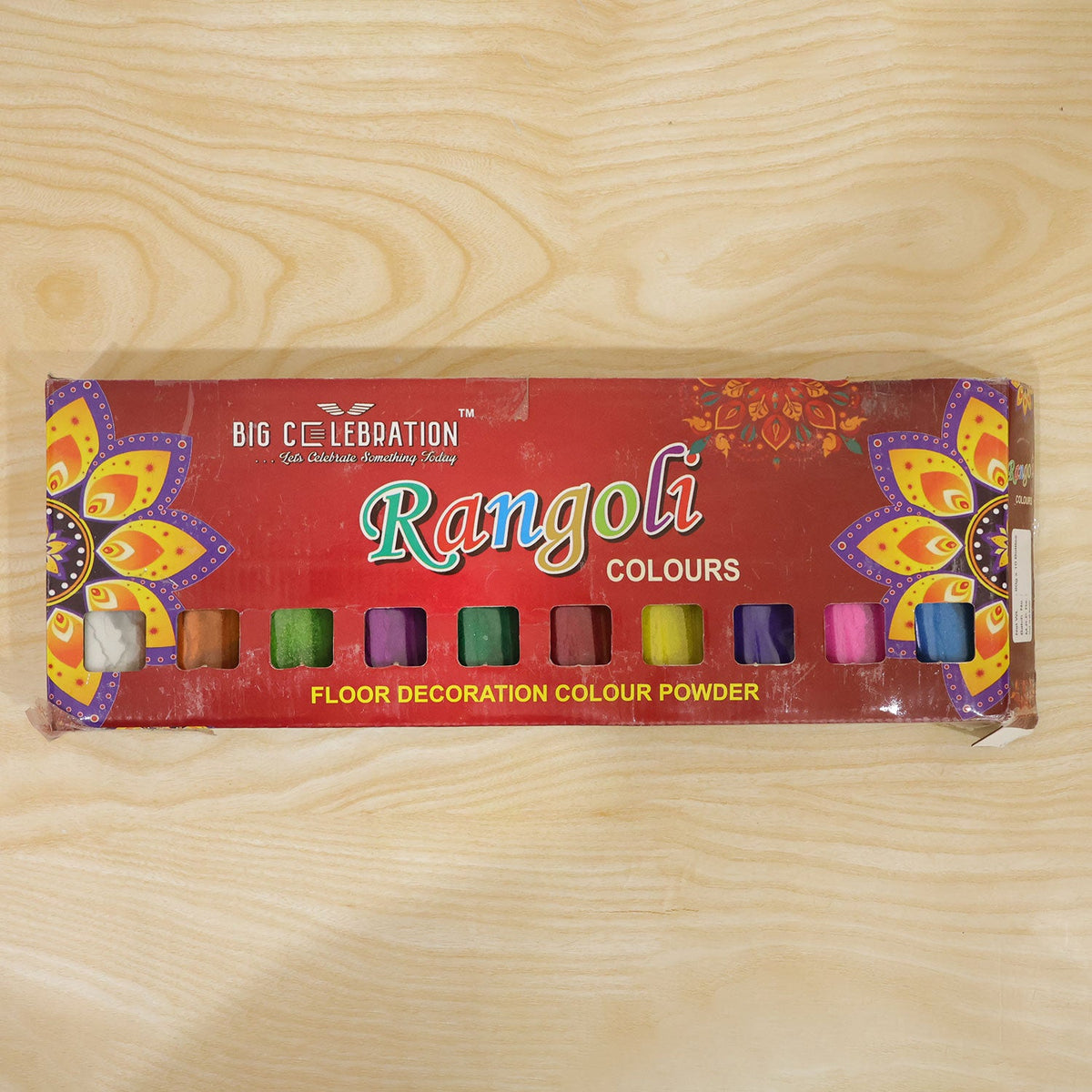 A red box of Rangoli colors, featuring ten small square windows revealing different powder colors, is displayed on a light
