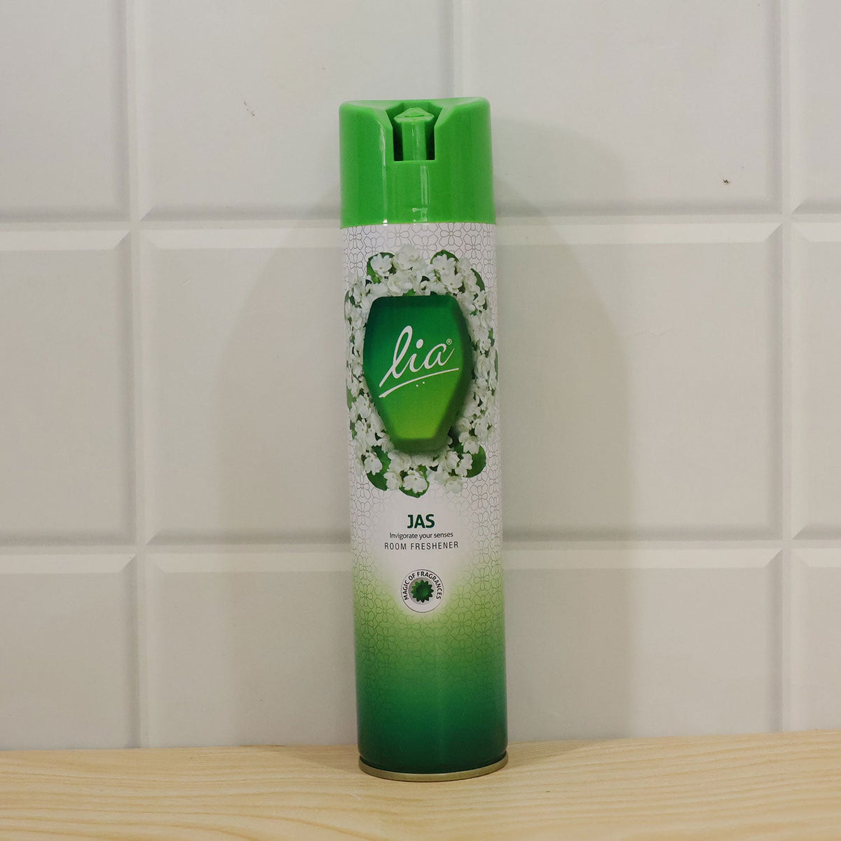 Green and white Lia Jas room freshener aerosol can on a wooden surface against a white tiled wall.