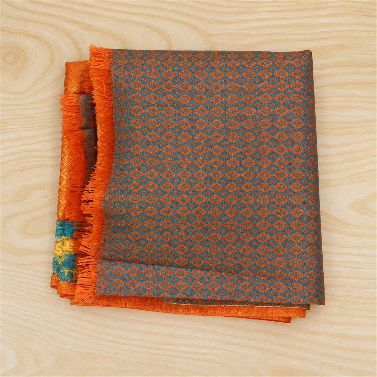 A folded orange and teal patterned shawl with fringe is displayed on a light wooden surface.