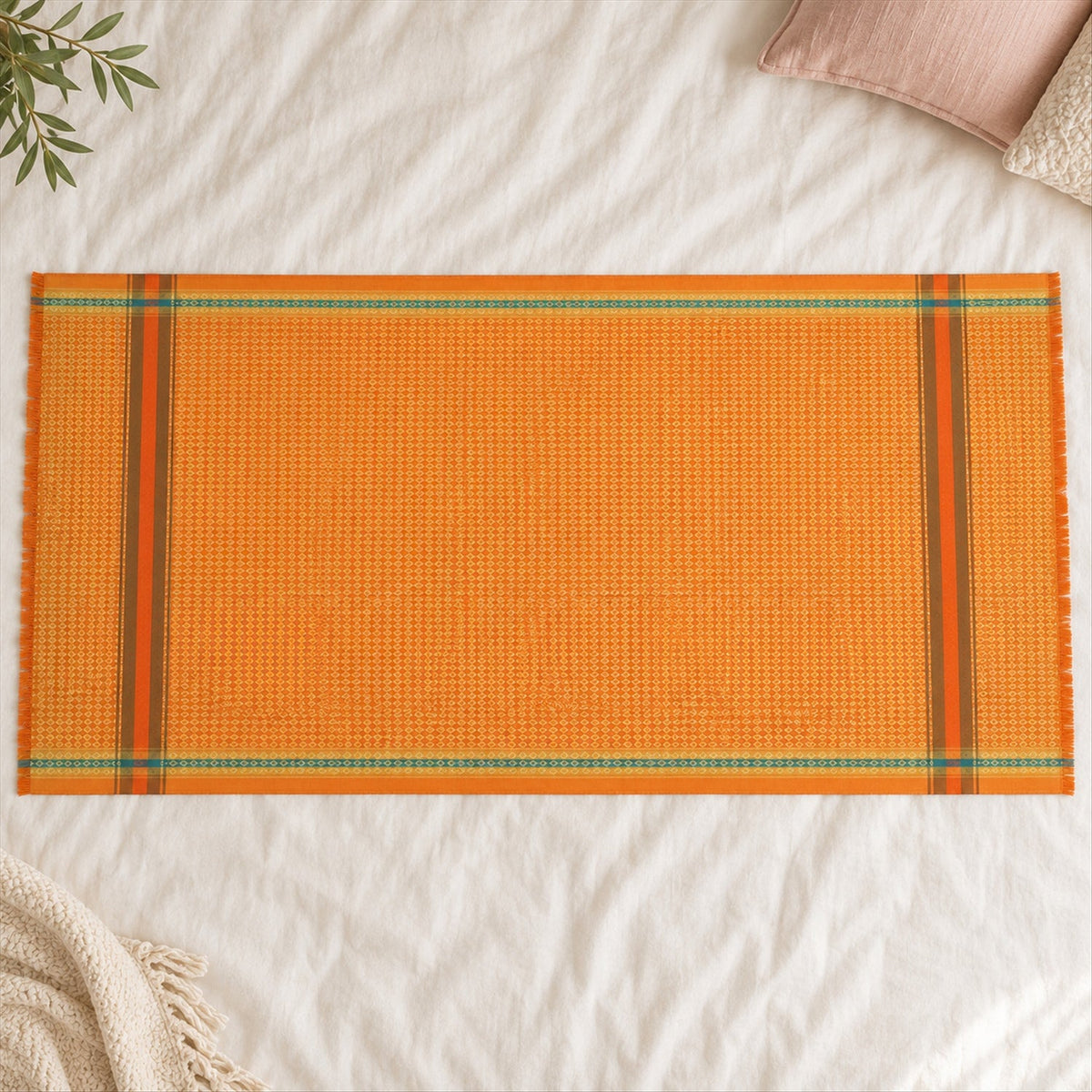 An orange shawl with diamond texture and striped ends is spread on a textured white blanket with pillows.