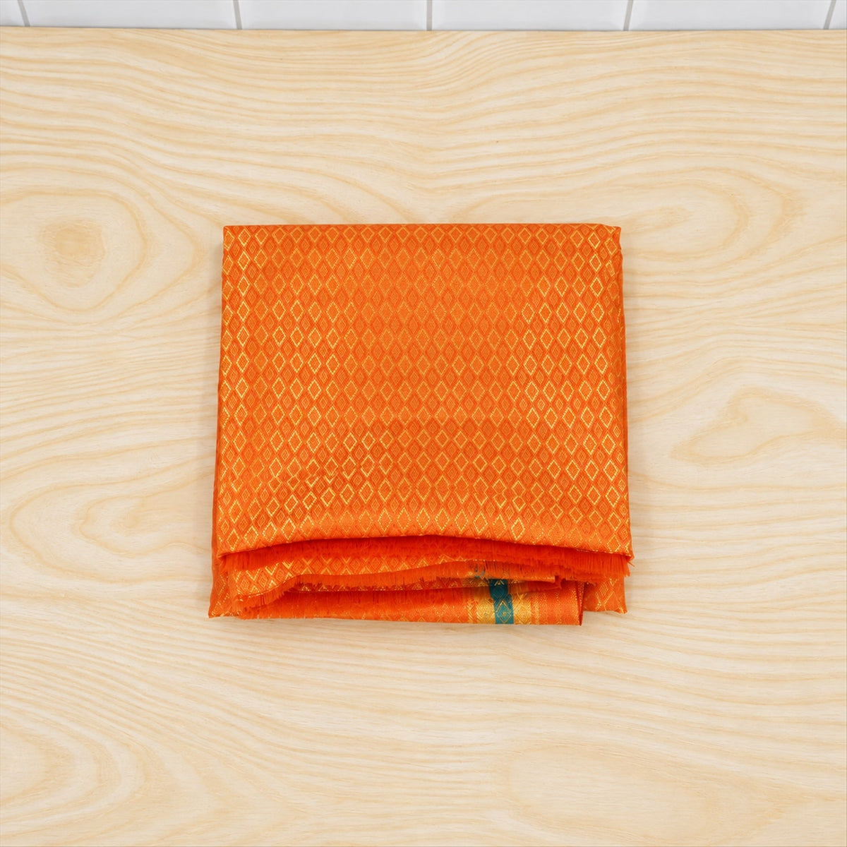 A neatly folded orange shawl with a diamond pattern and striped border rests on a light wooden surface.