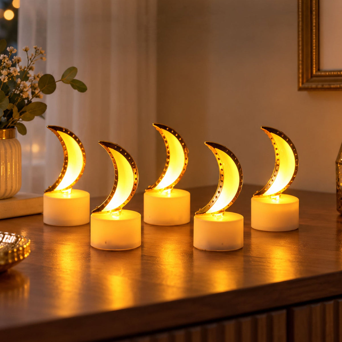 Five moon-shaped LED tealights with golden trim and warm glow, arranged on a dark wooden surface with a vase of flowers in