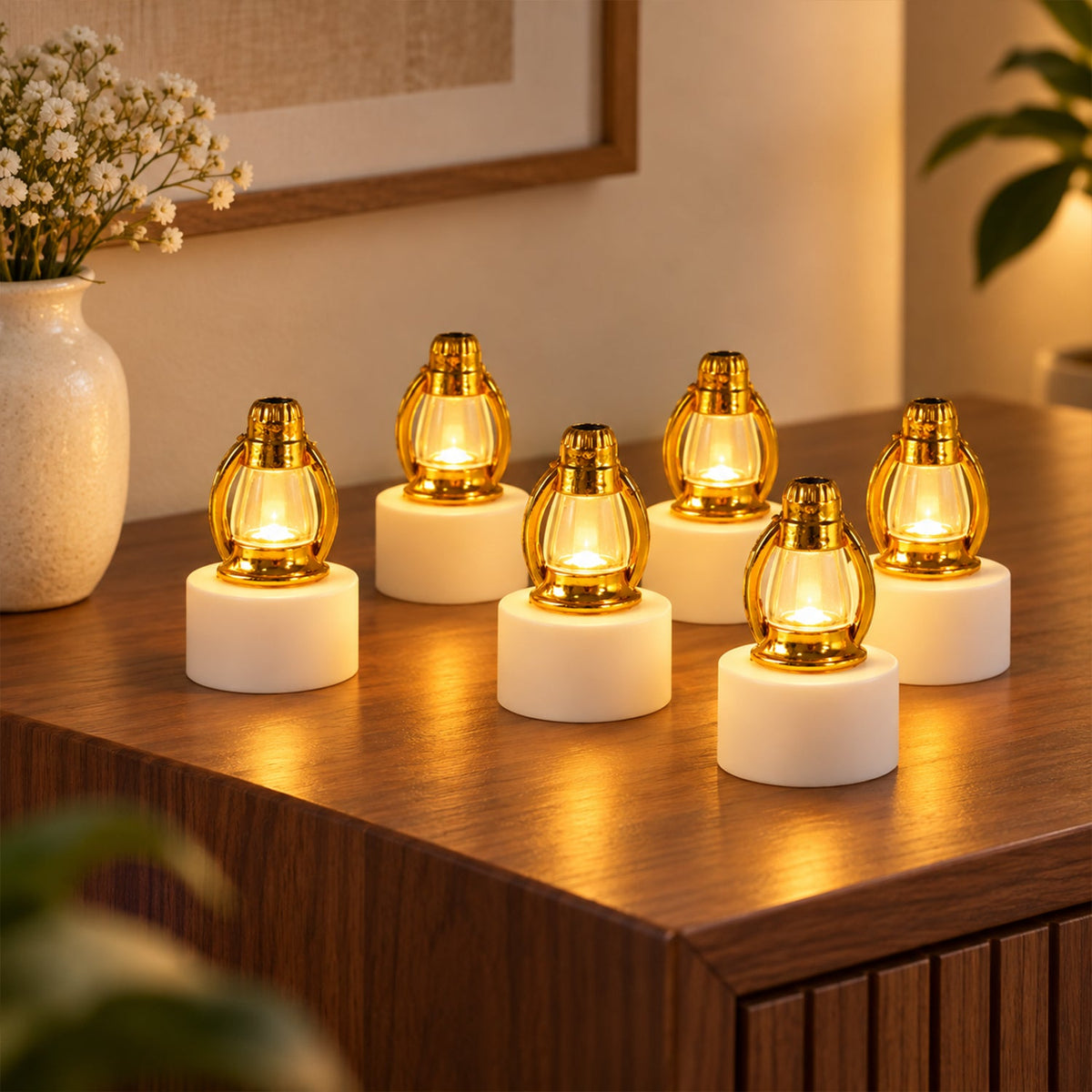 Six small gold and white LED lanterns glow with warm light on a wooden surface next to a vase of flowers.