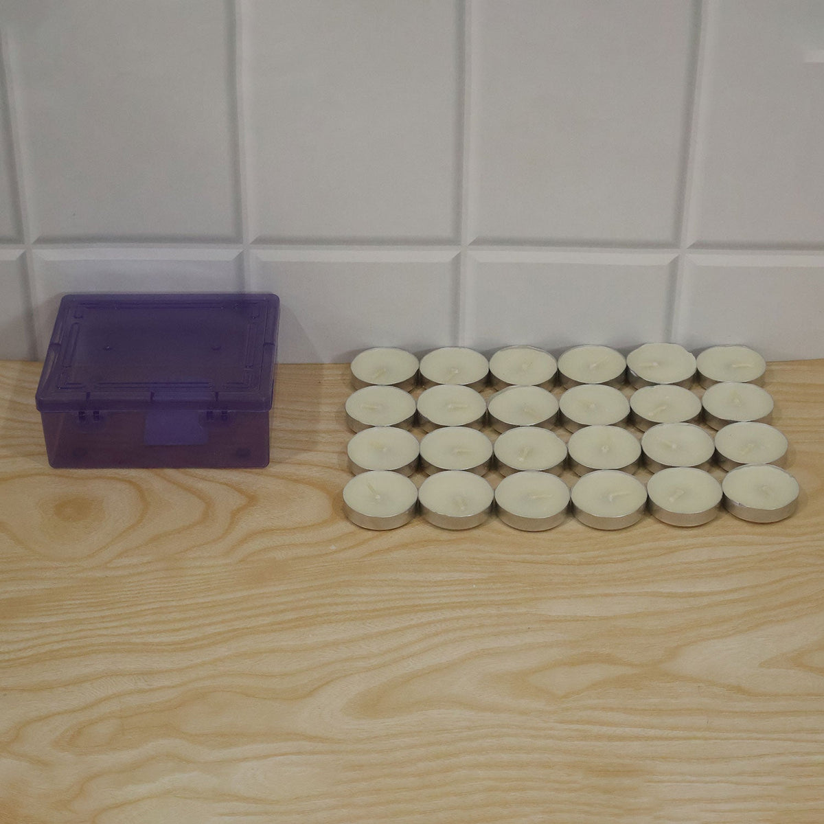 Twenty-four unlit white tealight candles next to a translucent purple storage box on a light wooden surface.
