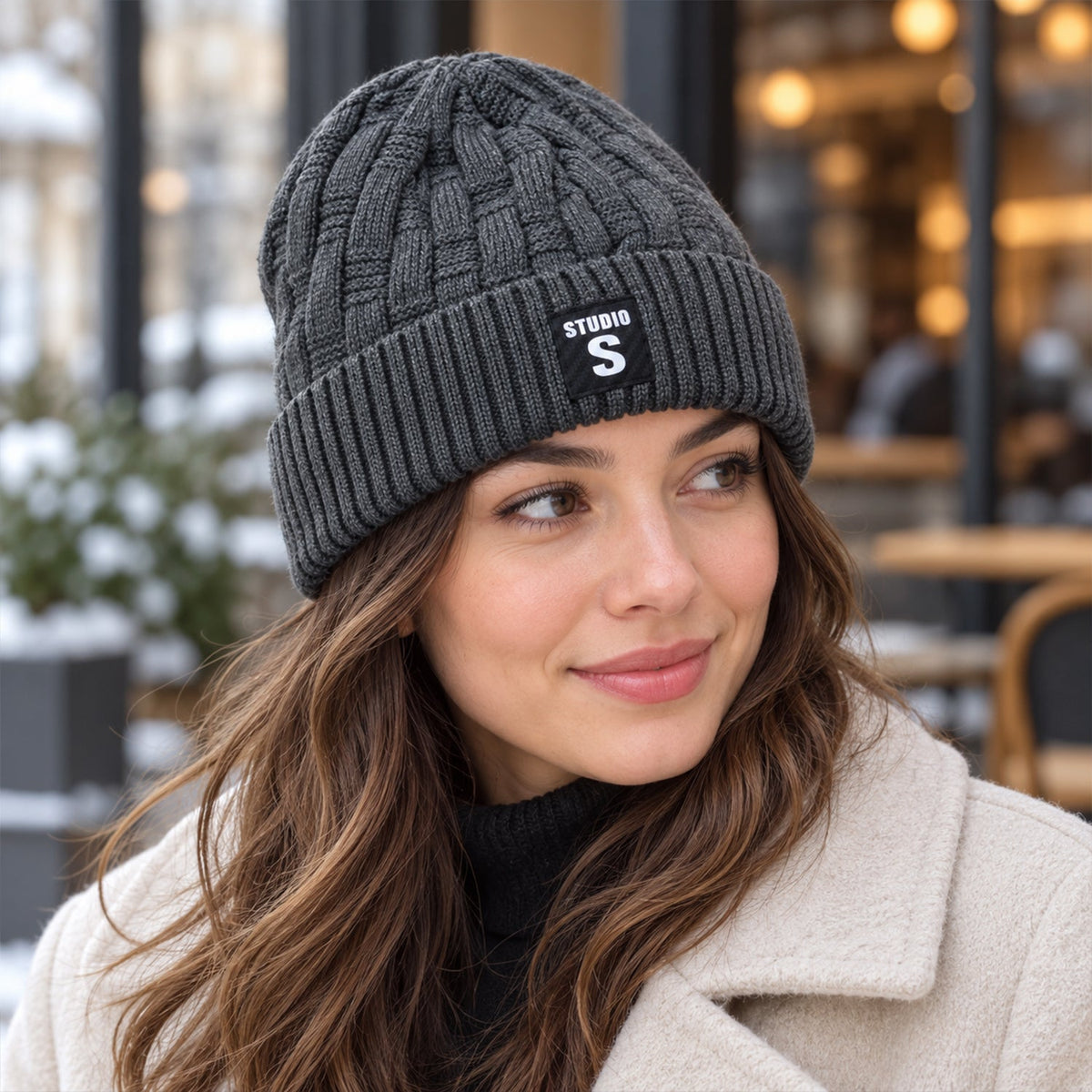 A woman wearing a dark grey cable knit beanie with a folded ribbed cuff and a black label, smiling outdoors in a snowy