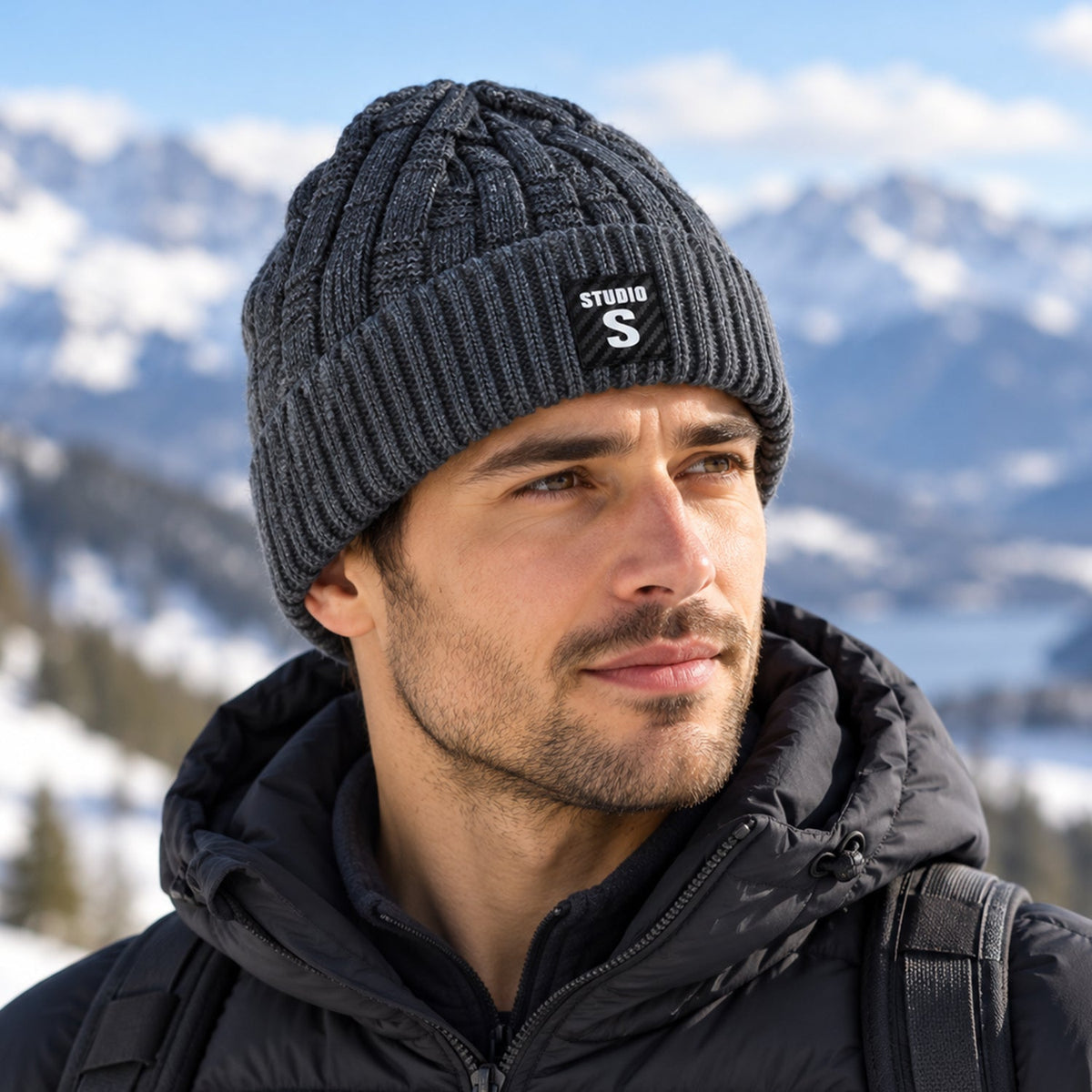A man wearing a dark grey cable knit beanie with a folded ribbed cuff and a black label, looking to the side in a snowy