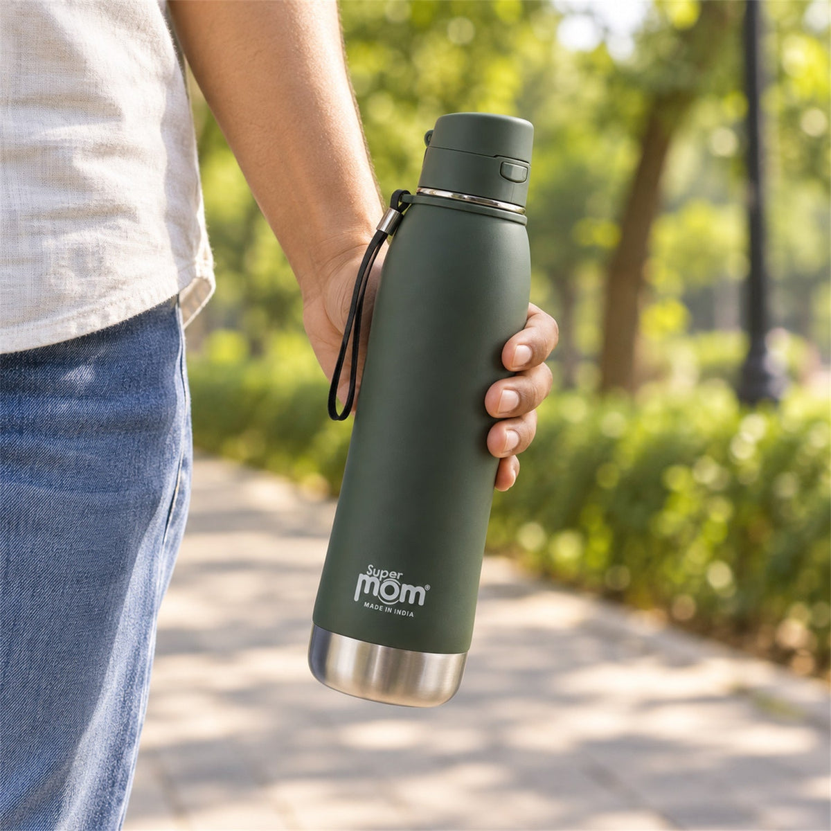 A person's hand holds a closed dark green insulated stainless steel water bottle with a wrist strap while walking outdoors.