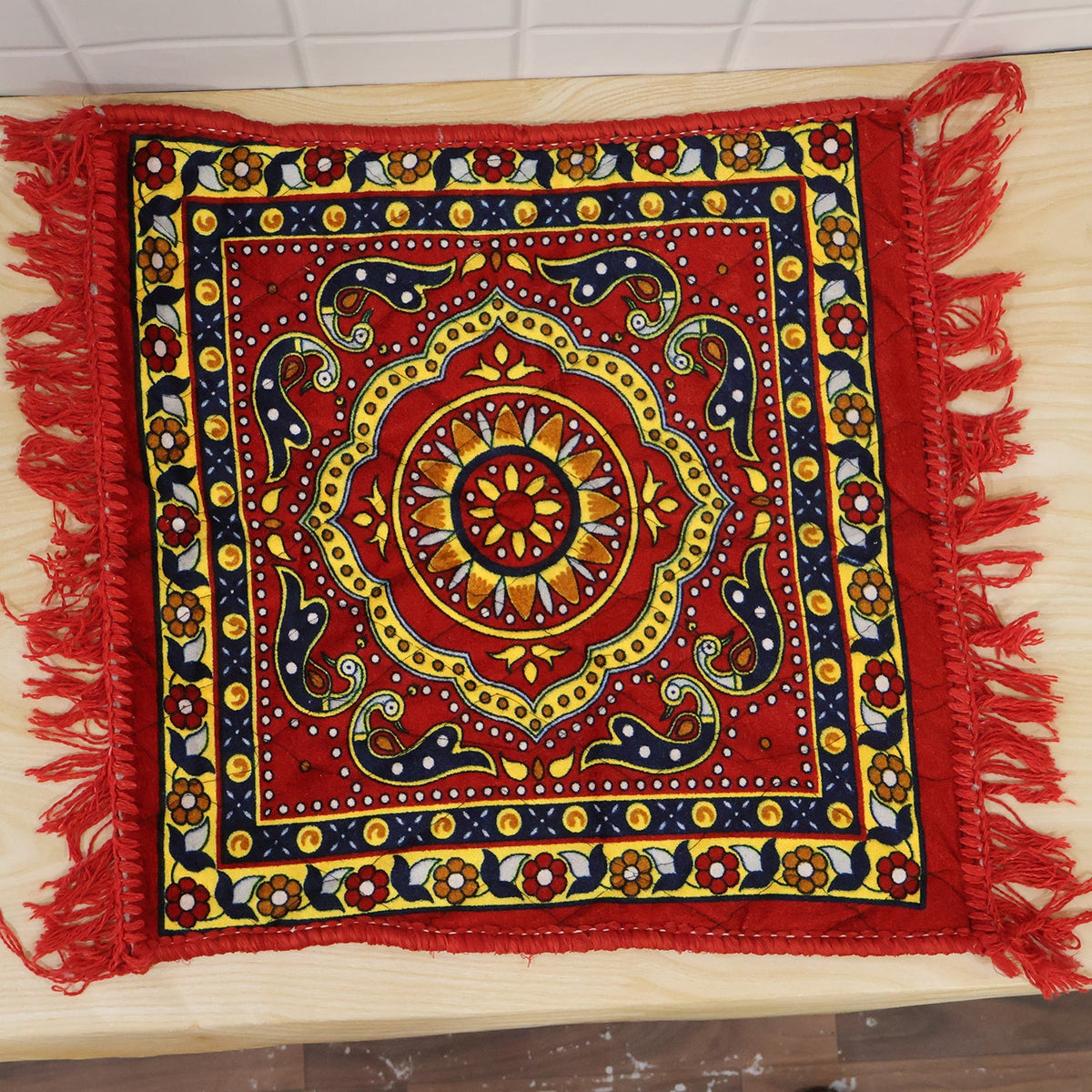 A red square prayer mat with a traditional yellow and blue pattern and red fringe, lying on a light wooden surface.