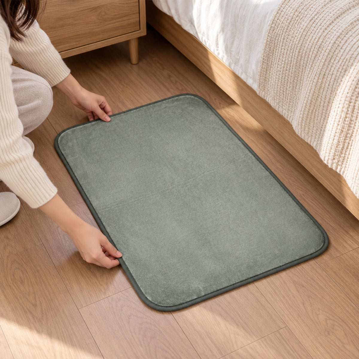 A person adjusting a grey, plush floor mat with dark trim on a wooden floor.
