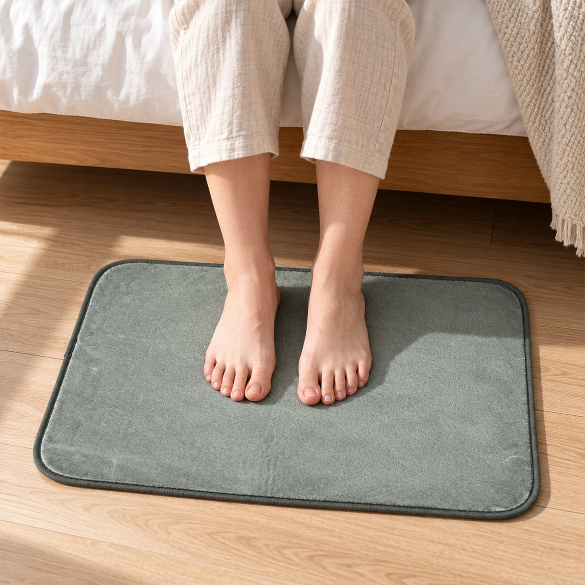 Bare feet standing on a soft, rectangular, grey floor mat next to a bed.