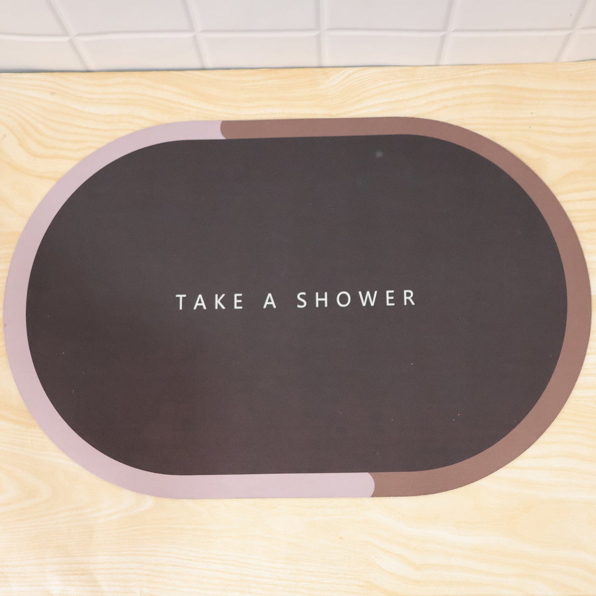 A dark oval bath mat with a pink and brown border and text is displayed flat on a light wooden surface.