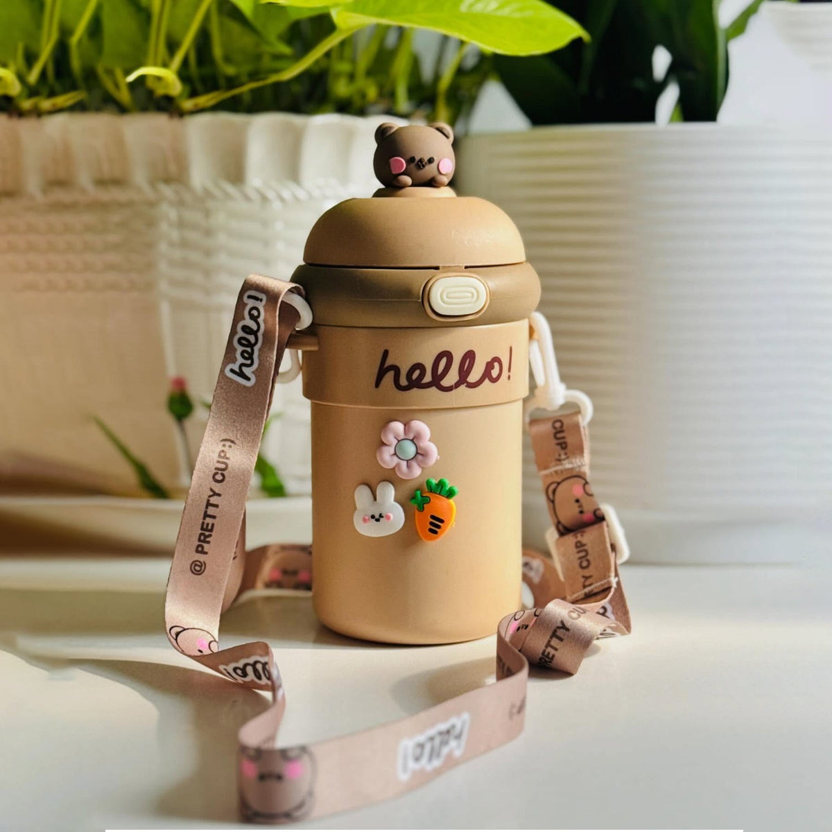 A tan sipper bottle with a bear on top, 'hello!' inscription, and cute animal and flower charms, with a patterned strap, on