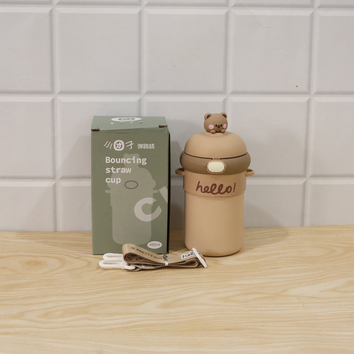 A tan sipper bottle with a bear on top, a patterned strap, and its product box, on a wooden surface.