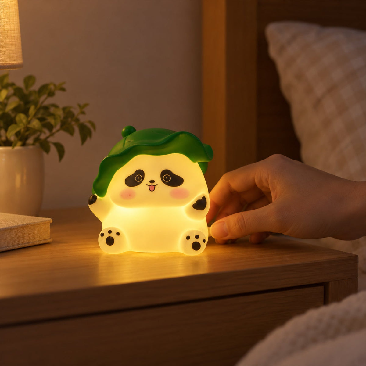 A hand reaches to touch the glowing panda night light on a bedside table in a dimly lit bedroom.