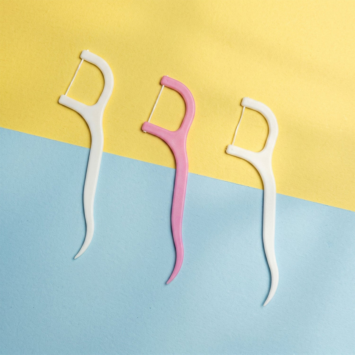 Three dental floss picks, two white and one pink, standing upright against a two-toned yellow and blue background.