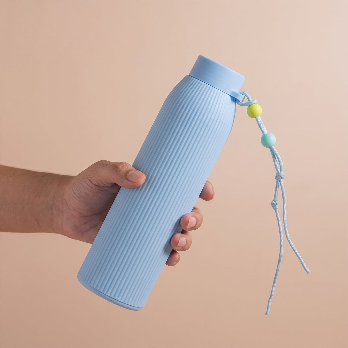 A hand holds a light blue ribbed glass water bottle with a strap adorned with yellow, blue, and green beads.