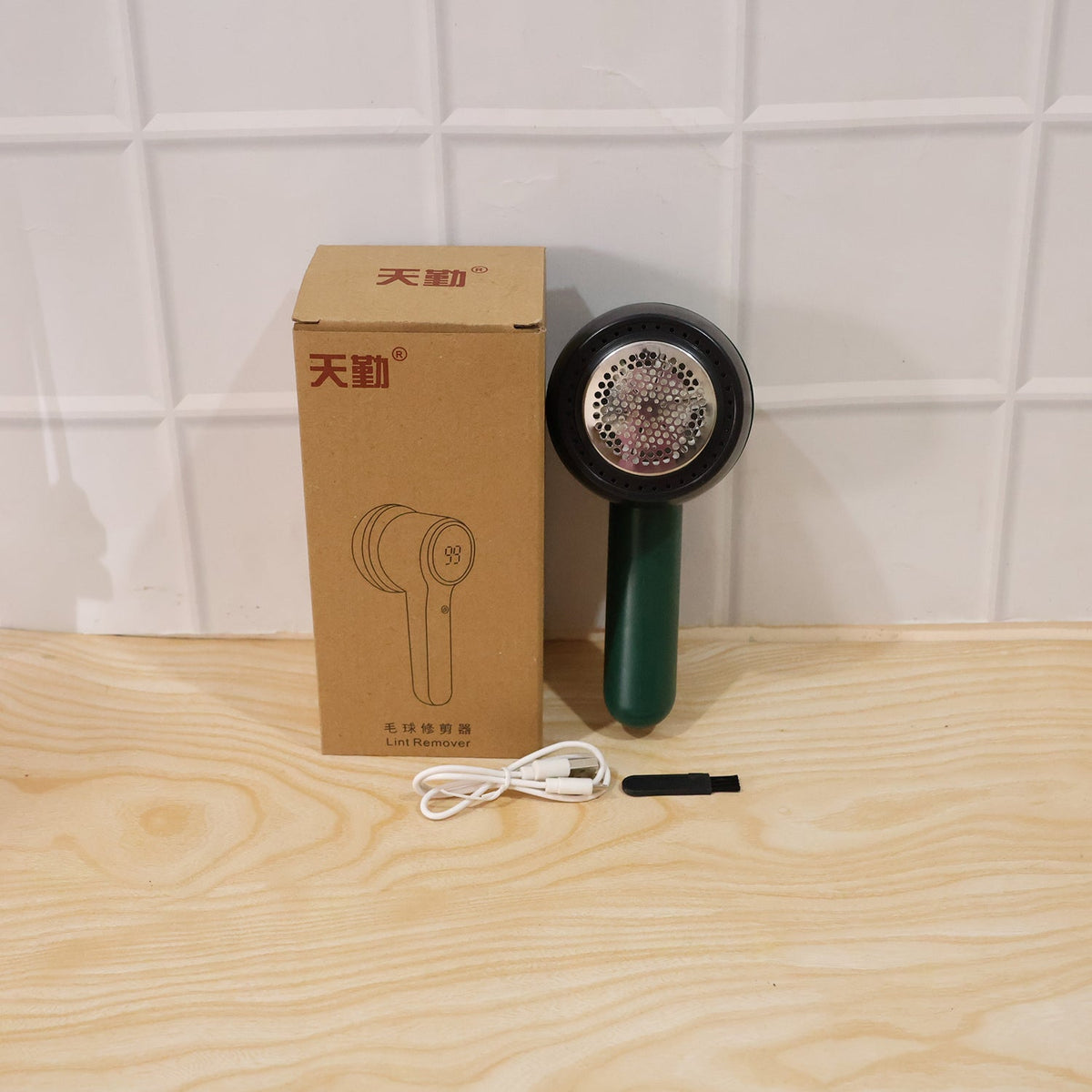A dark green and black lint remover, its brown box, a white USB-C cable, and a small black brush are arranged on a light