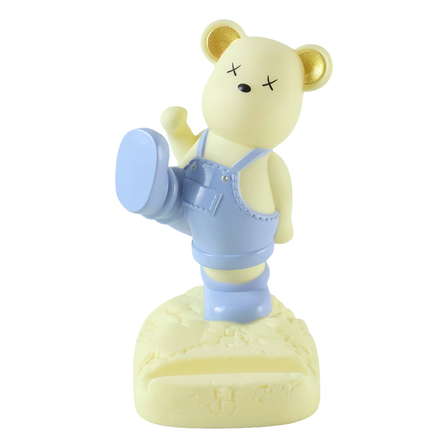A light yellow bear phone stand wearing blue overalls, standing on a textured base, against a white background.