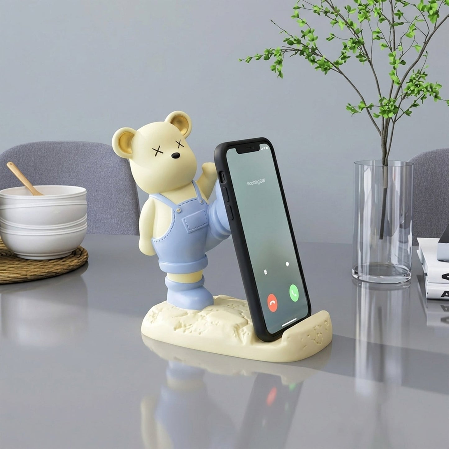 A light yellow bear phone stand holding a black smartphone vertically on a modern gray table with a plant.