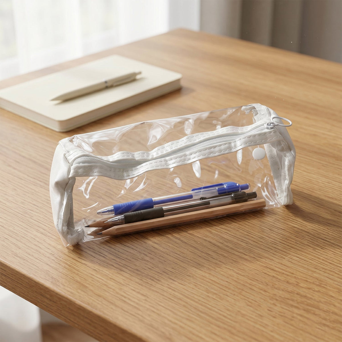 Clear PVC pencil case with white trim, containing blue pen and pencils, on a wooden desk with a notebook.