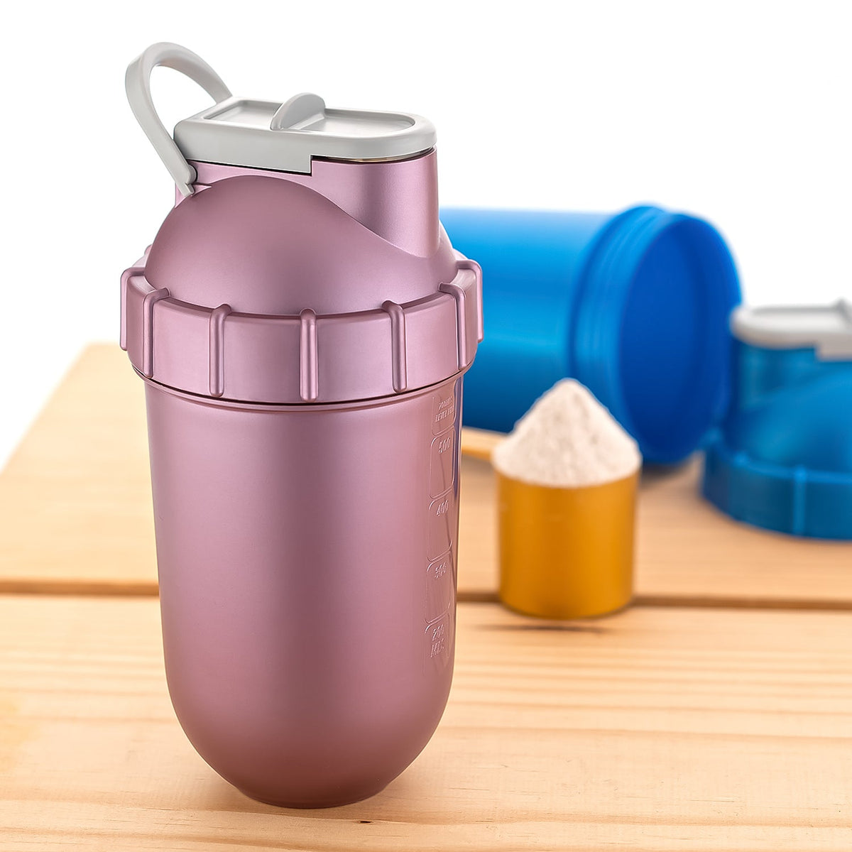 A pink shaker bottle stands on a wooden surface with a scoop of white powder and blue bottles in the background.