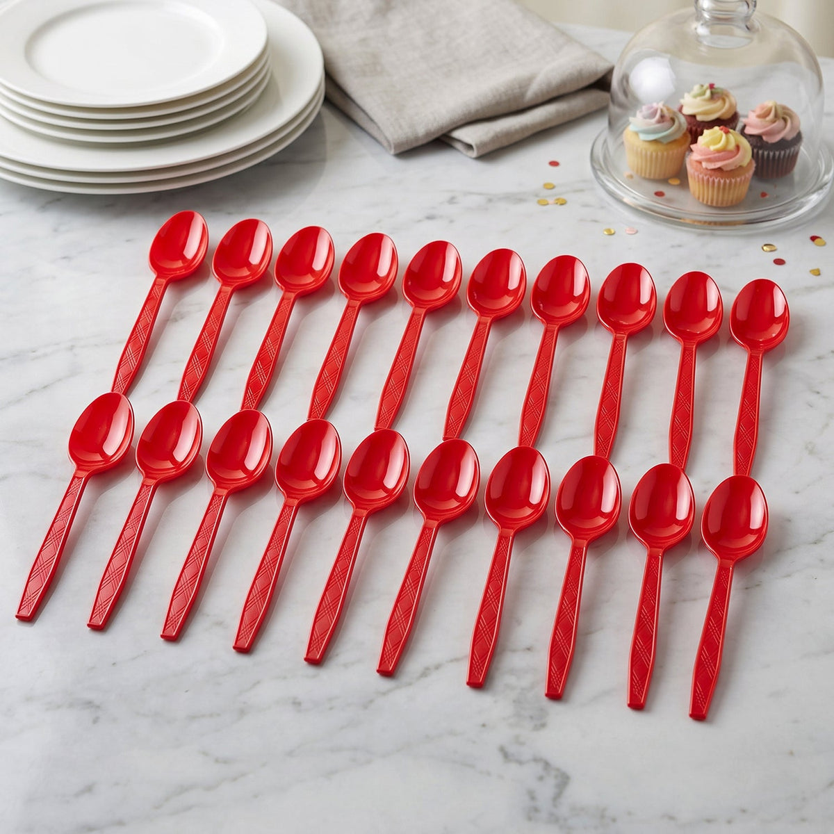 Twenty red plastic spoons arranged on a white marble table with plates and cupcakes in the background.