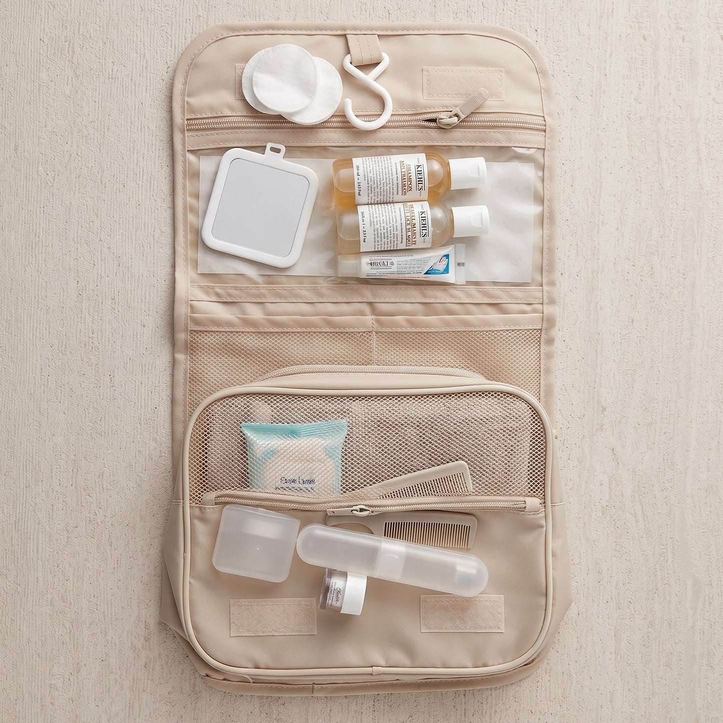 An open beige toiletry bag containing a mirror, small bottles, cotton pads, and a comb on a wooden surface.