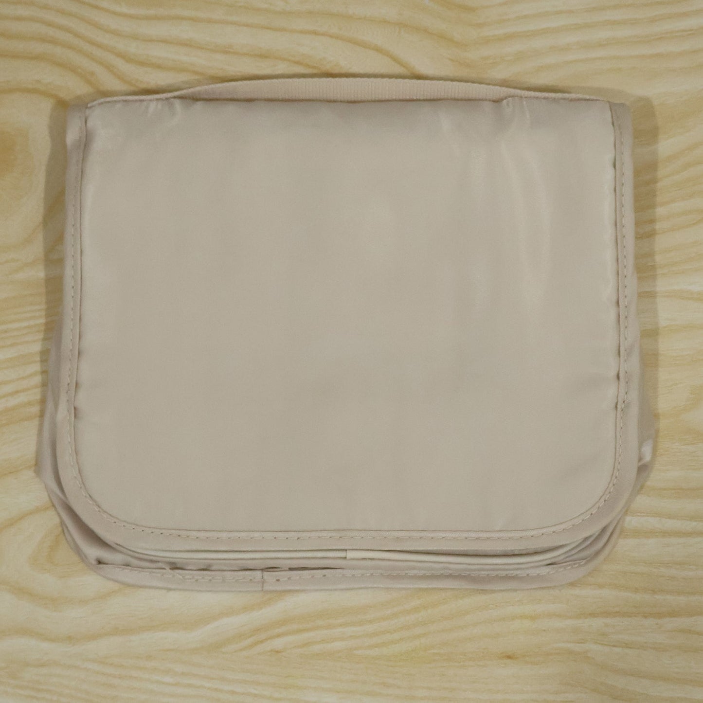 A neatly folded beige toiletry bag with a subtle sheen, resting on a light wooden background.
