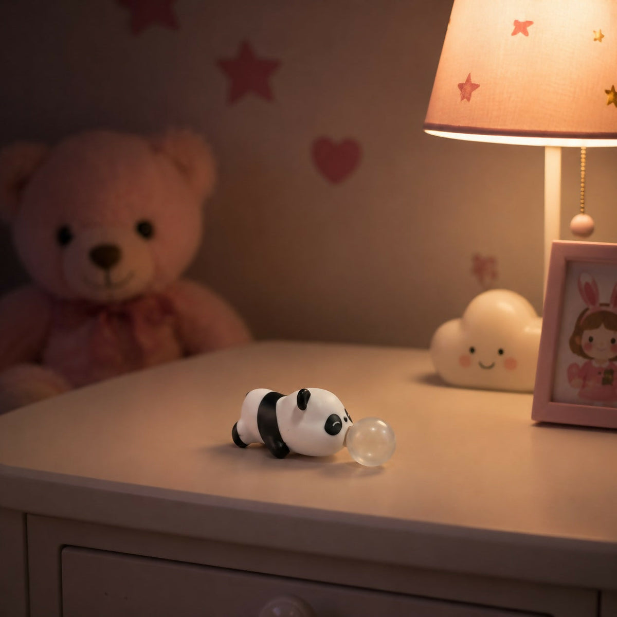 A small white and black panda toy with a clear bubble emerging from its mouth rests on a white nightstand next to a pink