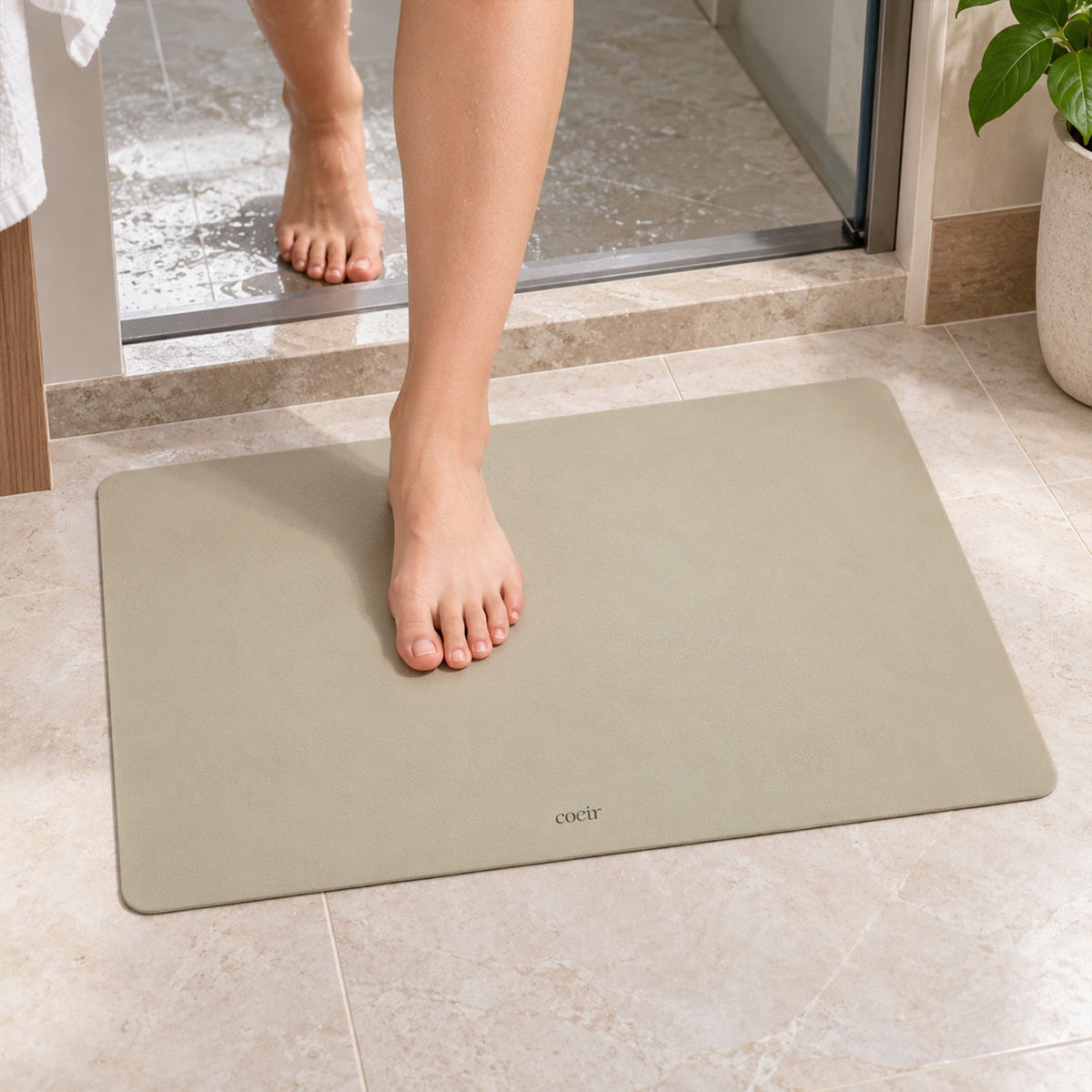 Bare feet step onto a light beige rectangular floor mat in a bathroom setting.