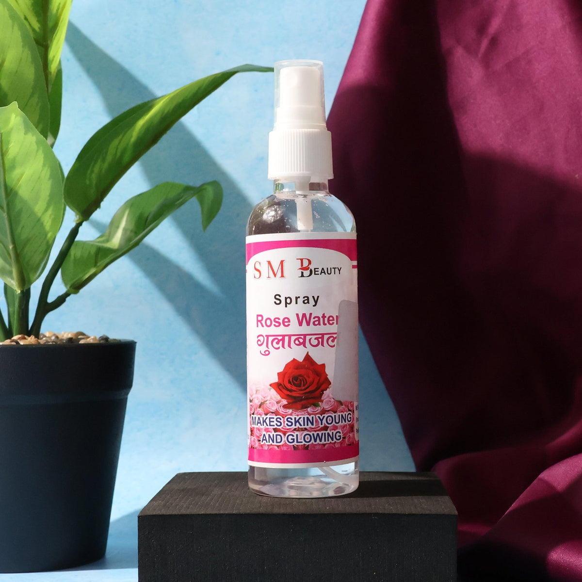 A clear bottle of SM Beauty rose water spray with a pink label sits on a black block next to a green plant.