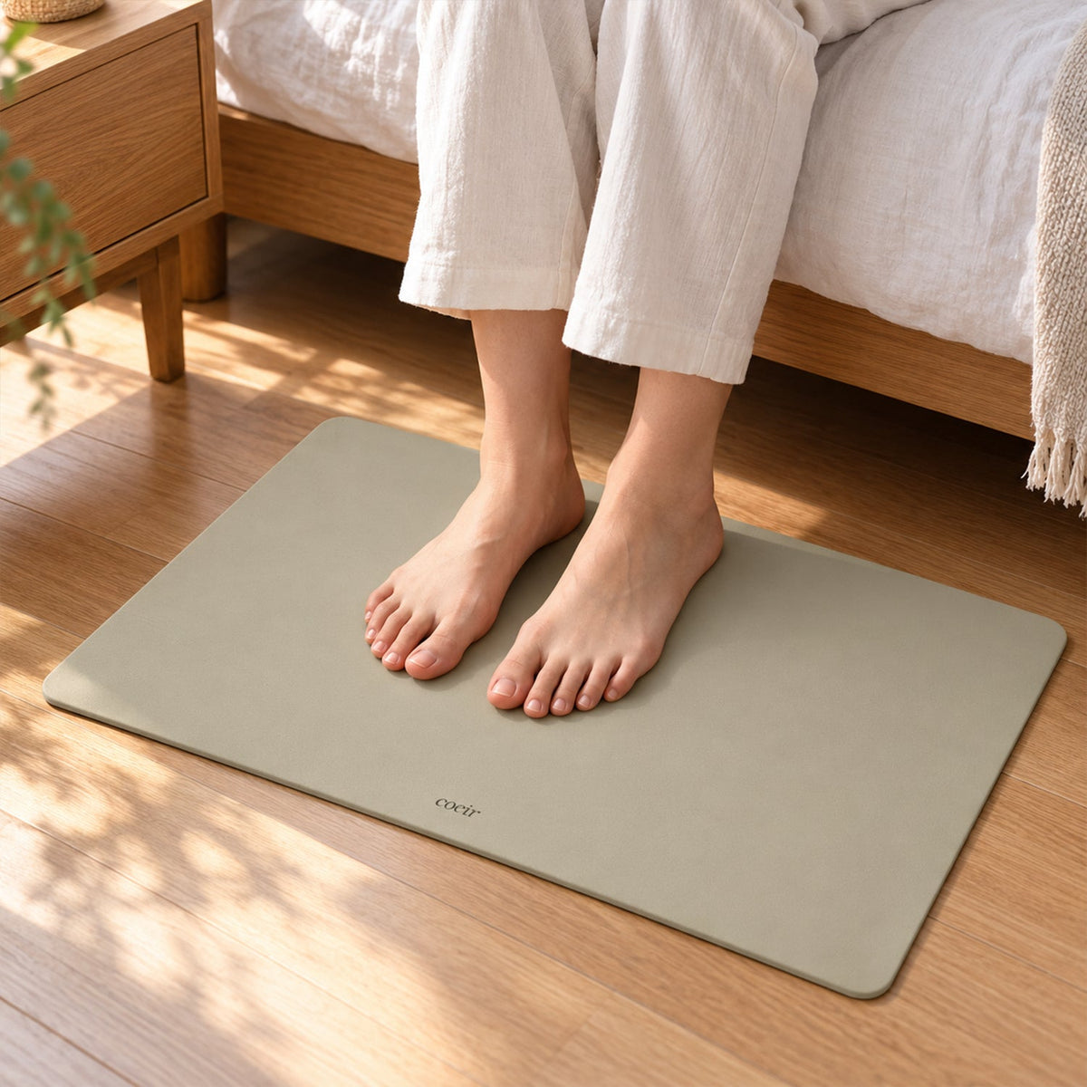 Bare feet are placed on a light beige rectangular floor mat next to a bed.