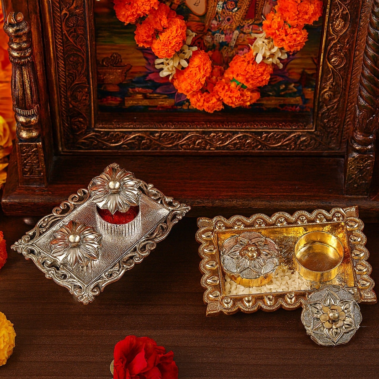 A silver and gold rectangular pooja thali with small containers, in front of a wooden altar with flowers.