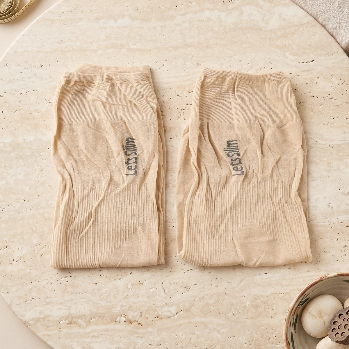Two folded beige arm sleeves with "Let's Slim" logo, on a light marble surface with decorative items.