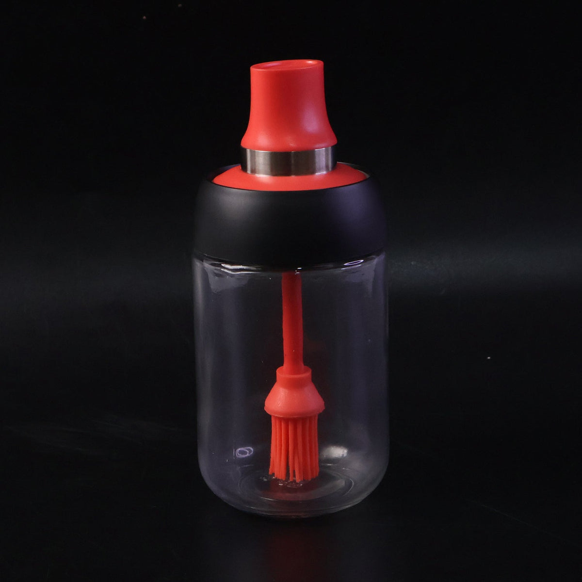 A glass oil dispenser with a red silicone brush and black top stands on a dark background.
