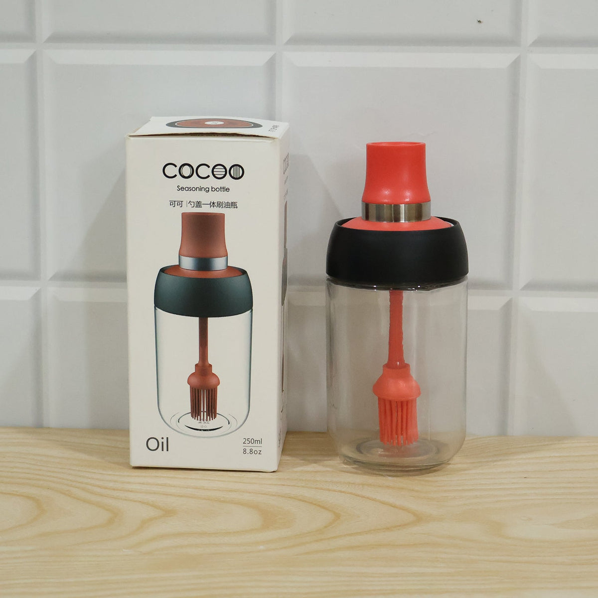 A glass oil dispenser with a red silicone brush sits next to its white and brown product box.