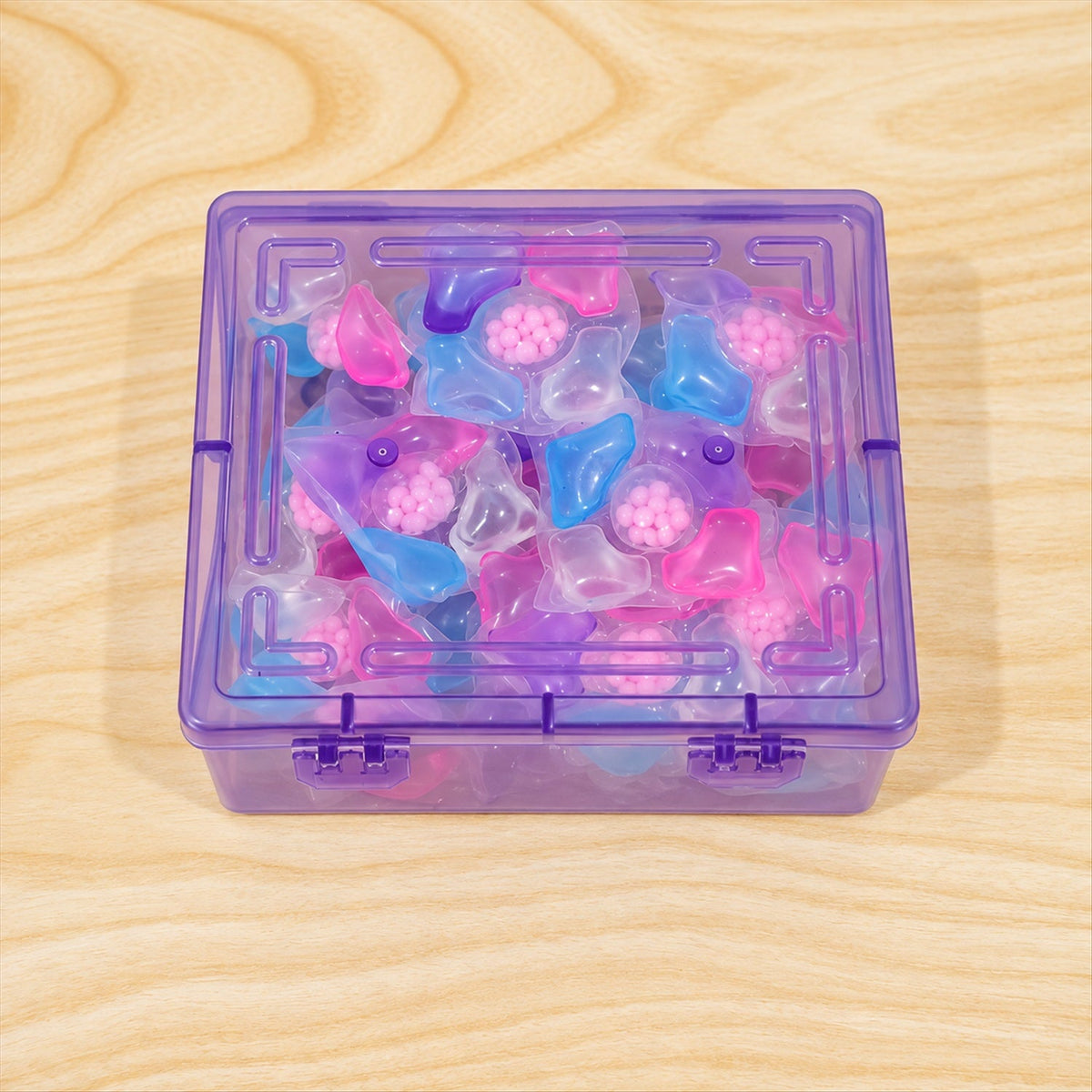 A clear purple plastic storage box, filled with an assortment of colorful laundry detergent pods, rests on a light wooden