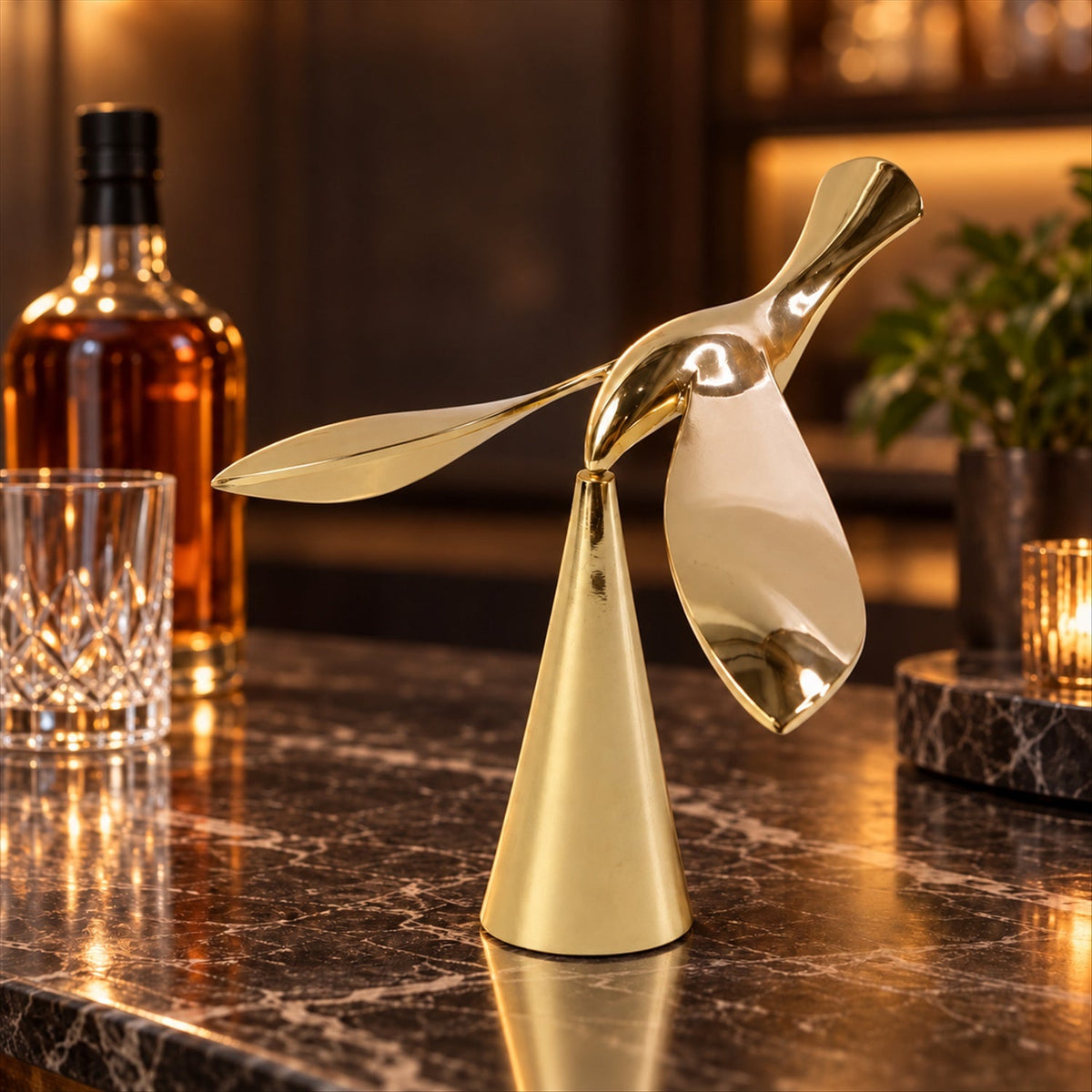 A gold balancing bird sculpture stands on a dark marble bar counter with a whiskey bottle and glass in the background.