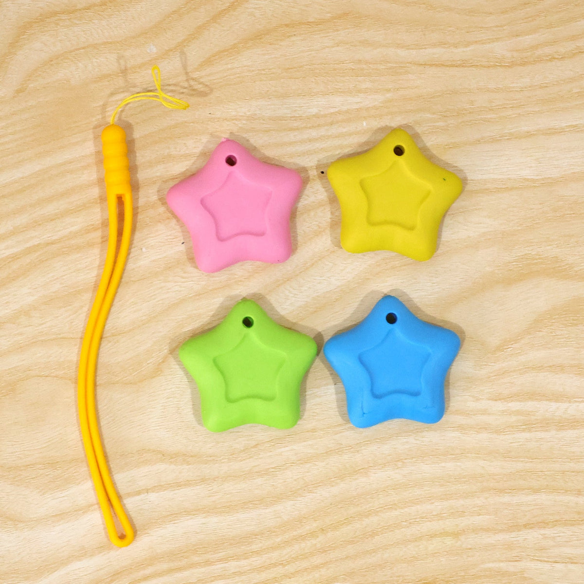 Four star-shaped erasers in pink, yellow, green, and blue are arranged on a light wooden surface next to a yellow lanyard.